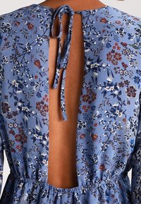 Vestido floral azul con espalda descubierta y cierre de lazo, que presenta una cintura fruncida y delicados patrones de flores en blanco, rojo y azul marino.