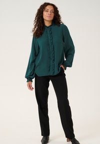 Teal blouse with ruffled front and cuffs, paired with black straight-leg trousers and pointed black ankle boots. Smooth fabric, relaxed fit.