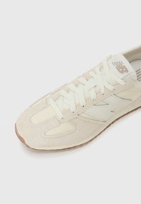 Beige idrettssko laget av semsket skinn og mesh-materiale, med hvite lisser og en logo på tungen, plassert mot en hvit bakgrunn.
