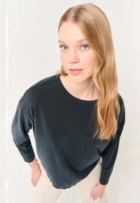 T-shirt nero a maniche lunghe con scollatura rotonda, texture morbida, vestibilità rilassata e spalle scese, abbinato a pantaloni di colore chiaro.