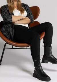 Botas negras de tobillo con suelas gruesas, pantalones negros y una camisa a rayas en blanco y negro, sentado en una silla marrón con un diseño curvado.