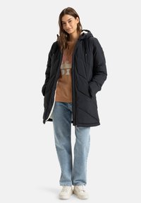 Svart dunjakke med hette, quiltet design, frontglidelås og sidelommer, brukt over en brun genser og lyseblå jeans.