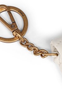 Μπρελόκ από μέταλλο σε χρώμα ορείχαλκου με συνδέσμους αλυσίδας, προσαρτημένο σε λευκό υφαντό δερμάτινο ύφασμα, με χαραγμένα τα γράμματα "PMKO" στο δαχτυλίδι.