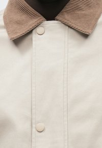 Μπεζ μπουφάν με υφασμάτινο καφέ γιακά από κορντόρι, που διαθέτει κεντρικό κούμπωμα με κουμπιά και ορατές ραφές.