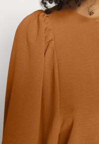 Top color ruggine con un tessuto morbido e testurizzato. Presenta dettagli arricciati sulle spalle e una silhouette ampia e fluida.