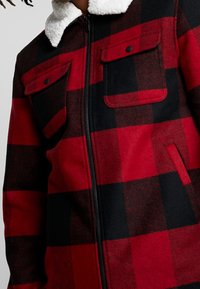 Punamusta ruudullinen takki, jossa valkoinen fleece-kaulus, vetoketjullinen etuosa, kaksi rintataskua napillisine läppineen ja sivutaskut, jota henkilö käyttää.