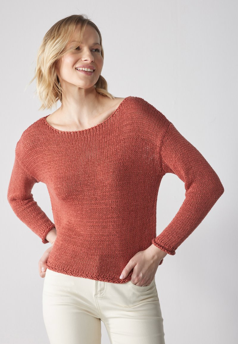 Maglione lavorato a maglia color ruggine con scollo ampio, maniche lunghe e un motivo testurizzato. Abbinato a pantaloni chiari.