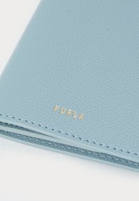 Lys blå teksturert lær lommebok med gull "Furla" merking; har synlige sømmer langs kantene. Kompakt og rektangulær design.