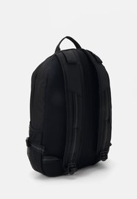 Mochila negra con diseño acolchado, paneles laterales de malla y cremalleras visibles. Cuenta con correas ajustables y forma curva para un soporte ergonómico.
