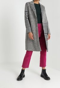 Grauer Hahnentrittmantel mit schwarz-weißen karierten Ärmeln, kombiniert mit rosa cropped Hosen und schwarzen geschnürten Stiefeln.