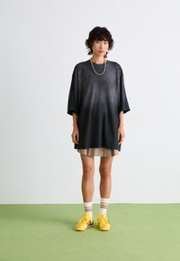 Oversized black satin shirt with short sleeves and a curved hem, layered over a beige skirt, paired with yellow sneakers and striped socks.