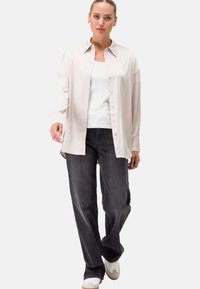 Camicia a maniche lunghe color beige chiaro con bottoni, canotta bianca, jeans grigio scuro e sneakers bianche. La camicia ha un colletto classico e una trama morbida.