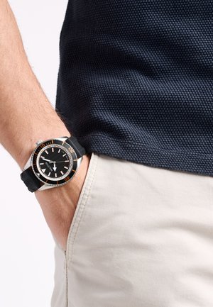 Black rubber strap watch with a silver case and black dial. Features orange accents and a textured fabric navy shirt, paired with beige pants.