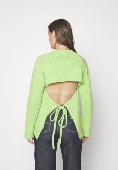 Pull en maille vert lime avec un dos ouvert, présentant un détail noué, des manches longues et des bords côtelés. Associé à un pantalon en denim foncé.