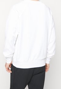 Person sedd bakifrån iklädd en enkel vit långärmad sweatshirt och mörka byxor mot en ljus bakgrund.