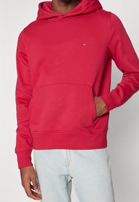 Hombre vestido con una sudadera con capucha roja y un pequeño logo en el pecho, jeans de un azul claro, con una mano en el bolsillo de la sudadera.