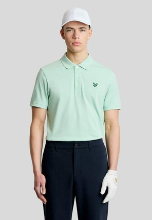 Ung mand iført en mintgrøn polo, navy bukser, hvid kasket og hvid golfhandske, står mod en ensfarvet lysegrå baggrund.