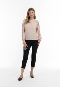 Lyserød bluse med samlet halsudskæring og lange ærmer, parret med sorte ankel-lange bukser og sorte flade sko på en neutral baggrund.