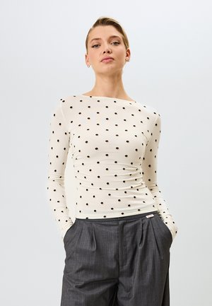 Hellbeiges Langarmshirt mit schwarzen Pünktchen, ausgestattet mit einem Bootsausschnitt und taillierter Form, kombiniert mit grauen plissierten Hosen.