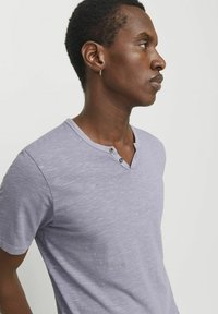 Camiseta de manga corta en color lavanda grisáceo con una textura suave, que presenta un escote en V y dos detalles de botones en la parte delantera.