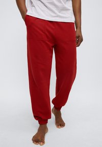 Κόκκινη φόρμα με άνετη εφαρμογή, κατασκευασμένη από απαλό ύφασμα. Διαθέτει δύο εμπρόσθιες τσέπες και ελαστικά τελειώματα. Λιτή υφή με μονόχρωμο χρώμα.