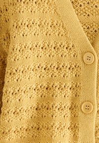 Cardigan jaune en maille avec motif ajouré semblable à de la dentelle et deux boutons ronds jaunes sur tissu texturé.