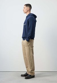 Joven de perfil vestido con sudadera azul marino con capucha, pantalones beige salpicados de pintura y zapatos negros sobre fondo liso.