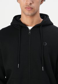 Ung mann iført en svart zip-up hettegenser med snorer og en liten subtil logo på venstre bryst.