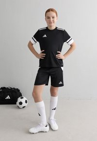 Svart fotbollskit med vita ränder, matchande shorts och långa vita strumpor. Har en logotyp på tröjan och skorna. Fotboll och väska i närheten.