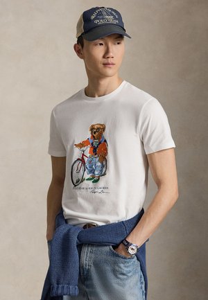 Fiatal férfi fehér pólóban, amin medve grafika van, kerékpáron, kék farmerral, tengerészkék sapkával és a derekán megkötött kék pulóverrel.