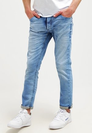 Mężczyzna w jasnoniebieskich podwiniętych dżinsach, białych sneakersach Reebok i białej koszulce z rękami w kieszeniach, stojący na jednolitym tle.