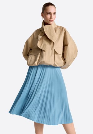 Kobieta w beżowej, oversize'owej kurtce z dużym kołnierzem i jasnoniebieskiej plisowanej sukience midi, stojąca na tle jednolitego tła.
