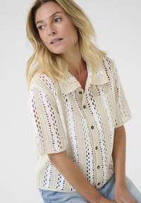 Crochet button-up shirt in cream with openwork pattern, short sleeves, and two chest pockets. Features light texture and wooden buttons.