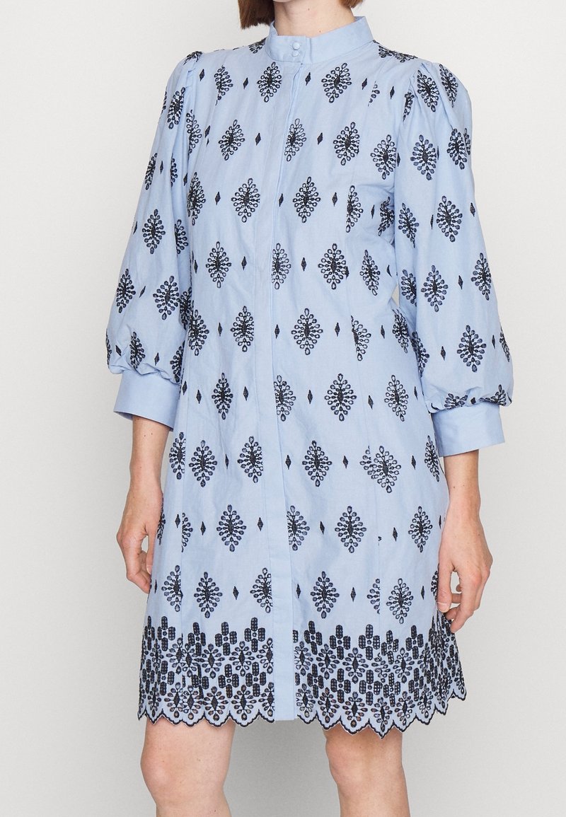 Jasnoniebieska sukienka koszulowa z czarnymi haftowanymi wzorami w kształcie diamentów, wysokim kołnierzem i bufiastymi rękawami, z falowanym dołem.