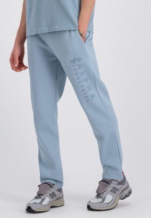 Blaue Jogginghose mit einem Kordelzugbund, dekoriert mit dem eingeprägten Text "ALPHA INDUSTRIES" entlang des linken Beins. Getragen mit grauen Sportschuhen.