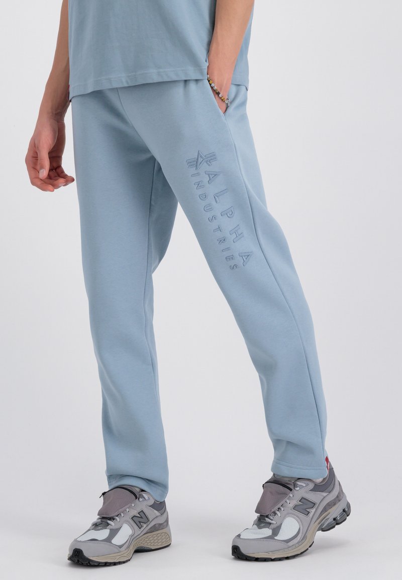 Blauwe sweatpants met een elastische tailleband, met de tekst "ALPHA INDUSTRIES" in reliëf langs het linkerpoot. Draagt met grijze sportschoenen.