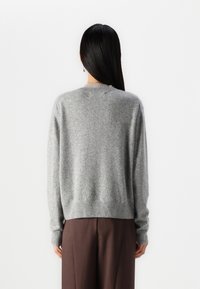 Femme aux longs cheveux foncés portant un pull en maille gris et un pantalon large marron, debout face à un fond blanc uni.