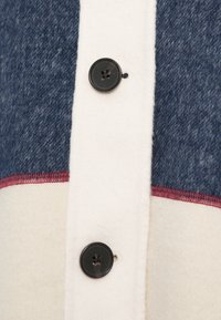 Πουκάμισο με κουμπιά, χαρακτηριστικό μπλε υφασμάτινο υλικό με υφή, μαλακό λευκό υλικό και κόκκινη λεπτομέρεια ραφής, με μαύρο κουμπί δύο τρυπών.