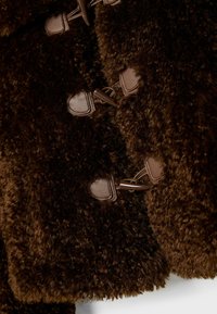 Tmavě hnědý kabát z umělé kudrnaté kožešiny s koženými lemovanými zapínáním na otočné klipsy v detailním záběru.