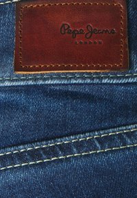 Tela de mezclilla azul con una etiqueta de cuero marrón que lleva grabado el texto "Pepe Jeans London". Costuras visibles en un color contrastante.