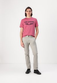 T-shirt rose à col rond avec le logo noir "PME LEGEND" imprimé, associé à un pantalon gris clair et des baskets noires, se tenant sur un fond blanc.