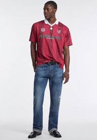 Camisa polo a rayas burdeos con cuello blanco, con logotipo y texto "GUESS JEANS". Llevada con jeans azules desgastados y zapatos negros.