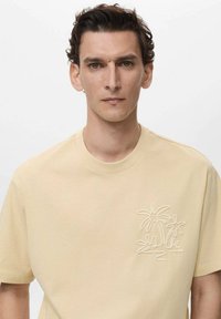 Tricou din bumbac de culoare galben deschis, cu mâneci scurte, având un design cusut cu palmier pe partea stângă. Textură moale, cu decolteu rotund.