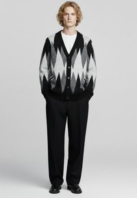 Cardigan nero e grigio con motivo a diamante, chiusura con bottoni e polsini a costine, abbinato a pantaloni neri e scarpe. Sotto, maglietta bianca semplice.