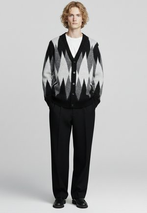 Cardigan nero e grigio con motivo a diamante, chiusura con bottoni e polsini a costine, abbinato a pantaloni neri e scarpe. Sotto, maglietta bianca semplice.