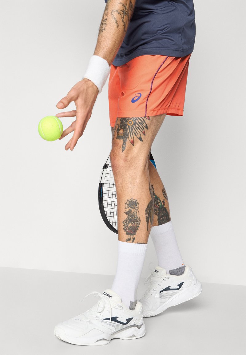 Orange sports shorts med blå detaljer, parret med hvide sneakers og ankelsokker. En grøn tennisbold holdes i den ene hånd.