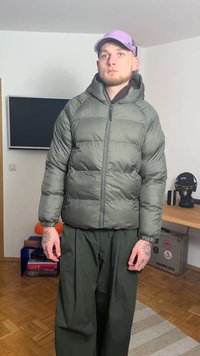 Groene pufferjas met een doorgestikte structuur, zwarte rits en capuchon. Wijdvallende groene broek. Lichtpaarse pet. Neutrale achtergrond met minimale decoratie.