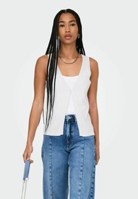 Cardigan bianco senza maniche con scollatura a V e chiusura a bottoni, abbinato a una canotta bianca e jeans blu a vita alta. Texture liscia, vestibilità casual.