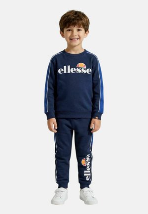 Garçon souriant debout portant un sweat-shirt bleu marine ellesse et un pantalon de jogging assorti avec des baskets blanches sur fond blanc.