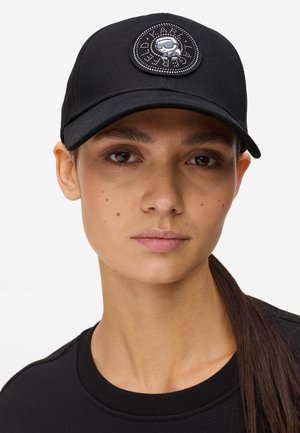 Γυναίκα με μακριά σκούρα μαλλιά που φοράει μαύρο καπέλο με στρογγυλό έμβλημα που απεικονίζει ένα κρανίο με γυαλιά ηλίου και το κείμενο "KARL".